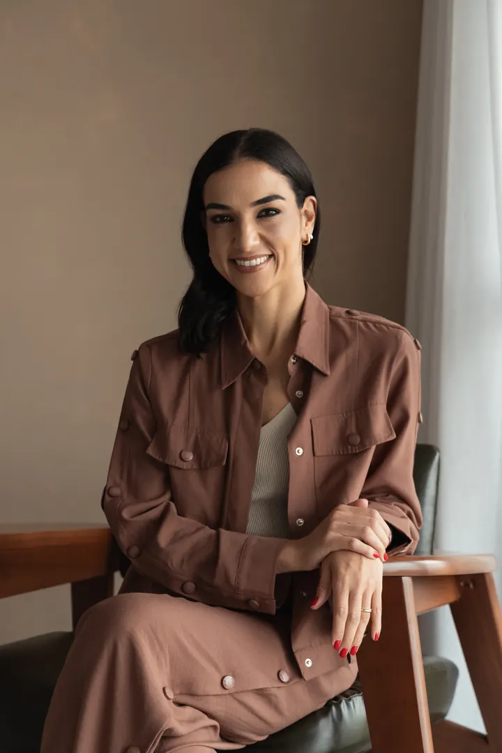 Dra. Luciana Paschoal, médica geriatra em São Paulo, sorrindo sentada em poltrona de madeira no consultório